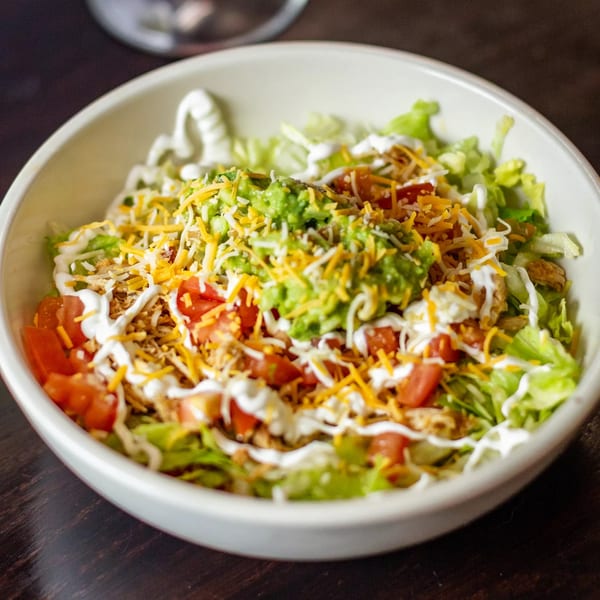 A vibrant Mexican salad with lettuce, tomatoes, cheese, guacamole, and sour cream in a white bowl.