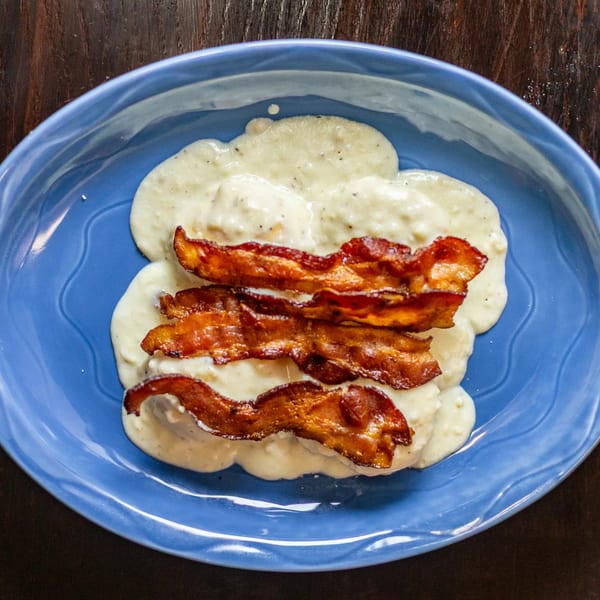 Biscuits and gravy topped with crispy bacon strips on a blue plate at Rudy's Little Hideaway.